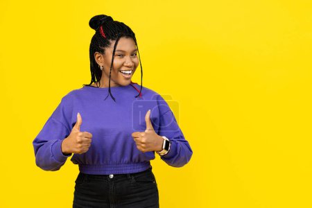 Photo for Afro woman giving a thumbs up gesture, dressed in casual purple attire against a vibrant yellow background. - Royalty Free Image