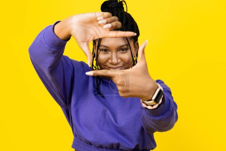 Foto de Mujer feliz enmarcando con los dedos en suéter púrpura sobre un fondo amarillo vibrante, expresando creatividad y diversión. - Imagen libre de derechos