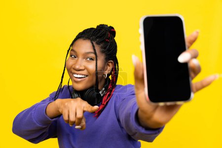 Téléchargez les photos : Femme pointant vers un écran vierge smartphone avec écouteurs sur un fond jaune vif. - en image libre de droit
