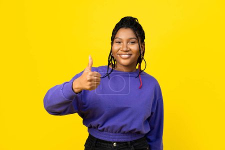 Foto de Mujer afro da un pulgar hacia arriba contra un fondo amarillo brillante usando un suéter púrpura, retratando positividad y aprobación. - Imagen libre de derechos