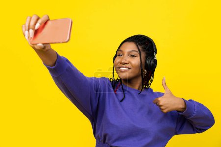 Foto de Mujer tomando una selfie con auriculares y dando un pulgar en suéter púrpura sobre fondo amarillo. - Imagen libre de derechos