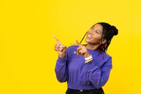 Téléchargez les photos : Jeune femme pointant vers le haut avec les deux mains sur un fond jaune vif dans un pull violet. - en image libre de droit