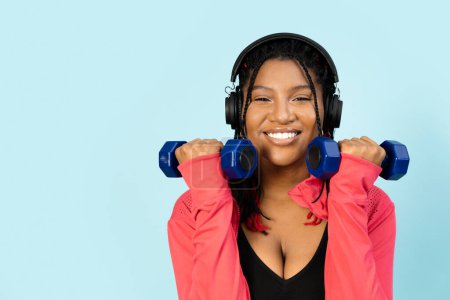 Photo for Woman working out with dumbbells while wearing headphones against a blue background.. - Royalty Free Image