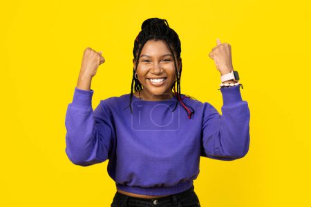 Foto de Mujer celebrando el éxito con una gran sonrisa, levantando los puños en una pose de victoria sobre un fondo amarillo brillante. - Imagen libre de derechos