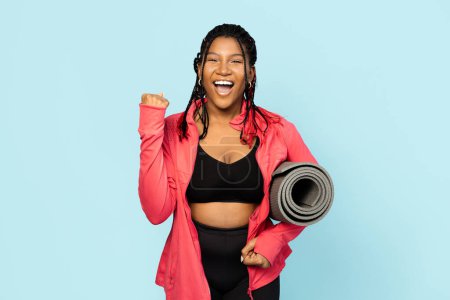 Photo for Woman ready for train in sportswear holding a yoga mat, celebrating fitness goals on a blue background. - Royalty Free Image