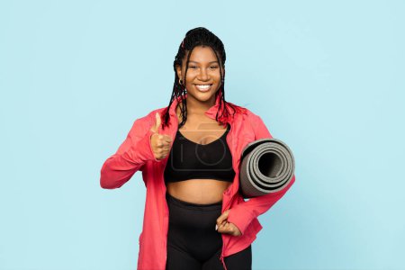Photo for Fitness Woman Giving a Thumb Up. Confident woman in sportswear with a yoga mat against a blue background, signaling readiness for a workout. - Royalty Free Image