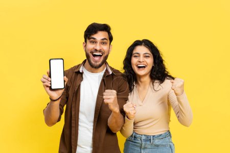 Téléchargez les photos : Une image vibrante d'un jeune couple joyeux gesticulant avec enthousiasme le succès tout en tenant un smartphone sur un fond jaune vif. Capture parfaite de l'émotion joyeuse, de la connectivité et du style de vie moderne. - en image libre de droit