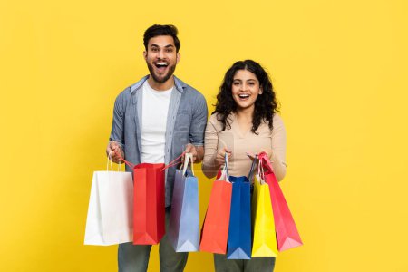 Téléchargez les photos : Un jeune couple joyeux souriant alors qu'ils tiennent une variété de sacs à provisions colorés sur un fond jaune vif, reflétant l'excitation du consumérisme au détail et des achats à la mode. - en image libre de droit