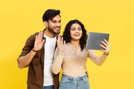 Téléchargez les photos : Les jeunes couples se rencontrent à l'aide d'une tablette, agitant et souriant sur un fond jaune vif, reflétant la joie de la communication numérique et la facilité de connexion en ligne. - en image libre de droit