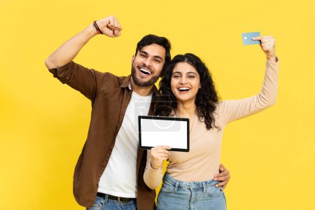 Téléchargez les photos : Couple excité avec carte de crédit et tablette numérique, achats en ligne, paiement, commerce électronique, fond jaune - en image libre de droit