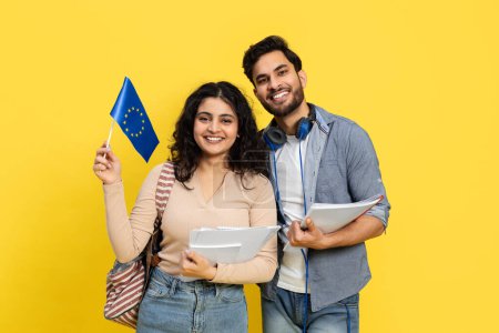 Téléchargez les photos : Joyeux jeunes étudiants tenant le drapeau de l'Union européenne avec des fournitures scolaires sur fond jaune - en image libre de droit