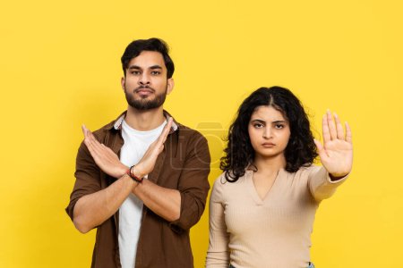 Young man and woman making stop gestures against a yellow background, expressing denial and boundaries with serious faces.