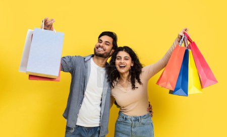 Couple heureux appréciant le shopping tenant de nombreux sacs de détail colorés sur un fond jaune vibrant. Capture parfaite de la diversité, de la mode et des concepts de consommation.
