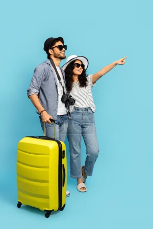 Photo for Couple On Vacation Pointing Away With Yellow Suitcase On Blue Background - Royalty Free Image