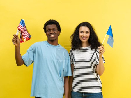 Diverse Freunde mit amerikanischen und europäischen Fahnen vor gelbem Hintergrund. Fröhliches multikulturelles Paar, Einheit, Freundschaft, Miteinander, Vielfalt, Austauschstudentenkonzept.