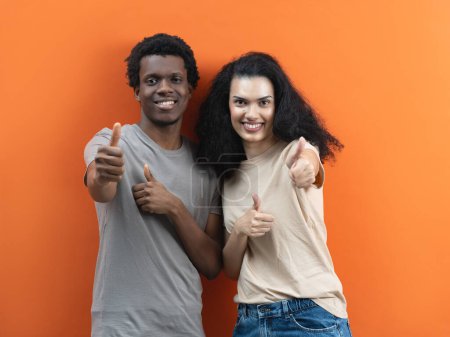 Photo for Happy Couple Giving Thumbs Up Sign, Young Adults Approval Gesture, Positive Feedback, Satisfaction Concept, Interracial Relationship, Joyful Expression, Casual Attire, Orange Background - Royalty Free Image