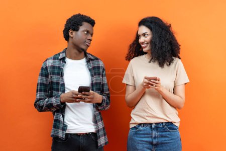 Téléchargez les photos : Jeune homme afro-américain et femme de race mixte utilisant des smartphones sur fond orange, médias sociaux, technologie, communication sans fil, mode de vie millénaire, connexion, personnes diverses - en image libre de droit