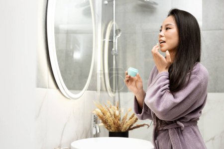Téléchargez les photos : Jeune femme appliquant crème visage pour la routine de soins de la peau dans une salle de bain moderne avec lumière naturelle. Beauté féminine et bien-être à la maison Concept. Expérience luxueuse de spa à domicile et rituel d'auto-soin. - en image libre de droit