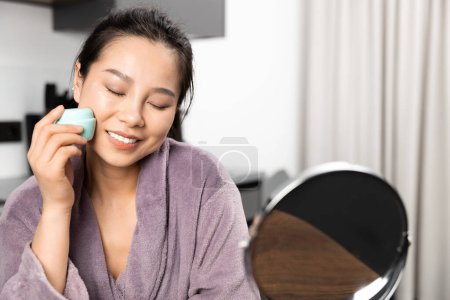 Photo for Radiant Young Woman Enjoying Skincare Ritual. Smiling Asian Lady In Bathrobe Applies Facial Cream For Beauty Routine. Self-Care And Wellness Concept With Mirror In Background. - Royalty Free Image