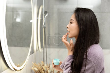 Téléchargez les photos : Femme appliquant le baume à lèvres dans le miroir de salle de bains Réflexion Self-Care Concept d'hygiène de routine - en image libre de droit