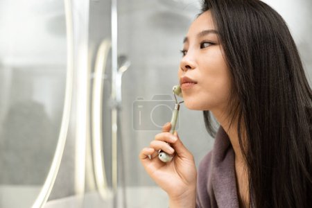 Mujer usando rodillo de jade en la cara para rutina de cuidado de la piel. Una joven mujer se involucra en un proceso de autocuidado, deleitándose en un ritual de belleza y bienestar con un rollo de piedra de jade natural.