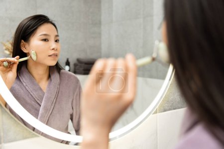 Young Woman Using Jade Roller For Beauty Routine. Young Asian Female In Bathrobe Performing Skincare Treatment With Facial Roller, Beauty Concept, Self-Care, Spa Day At Home. 