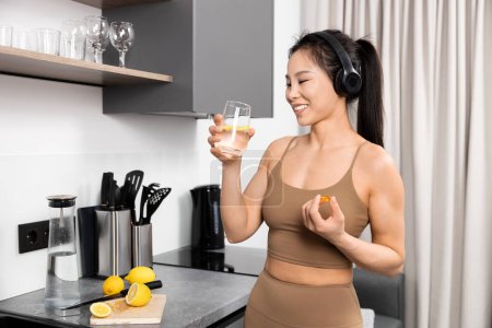 Lächelnde Frau in Sportbekleidung mit Kopfhörern, die frisches Zitronenwasser in der Küche genießt. Wellness, Gesundheit und Hydrationskonzept.