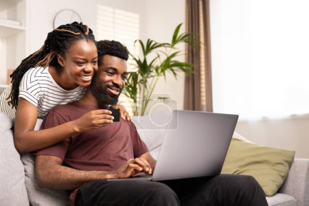 Téléchargez les photos : Joyeux couple afro-américain utilisant une carte de crédit pour magasiner en ligne, assis confortablement sur le canapé à la maison. - en image libre de droit