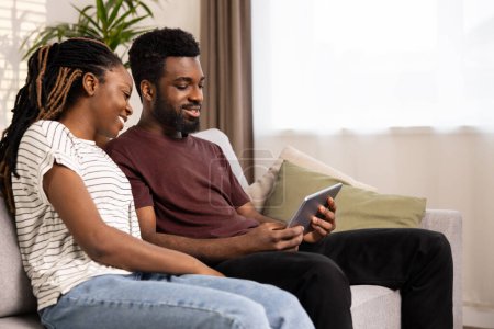 Photo for Smiling Couple Enjoying Digital Tablet On Sofa. A Happy Afro Couple Relaxing Together On The Couch With A Tablet In A Cozy Living Room Atmosphere, Embodying Togetherness And Digital Lifestyle. - Royalty Free Image