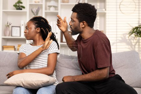 Konfliktlösung zwischen Paar zu Hause, Gefühle, Missverständnisse, Afro-Mann und -Frau sitzen auf Couch, Mann versucht sich zu entschuldigen oder mit Gesten zu erklären, während Frau wütend oder aufgebracht aussieht.