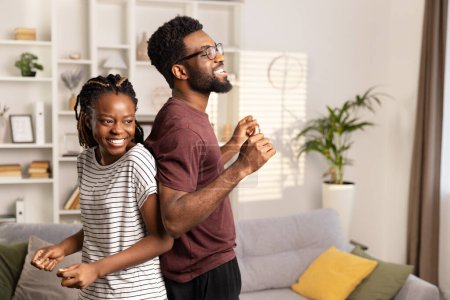 Foto de Joven pareja alegre compartiendo un baile en su acogedora sala de estar, irradiando energía positiva y unión en un entorno casual. - Imagen libre de derechos