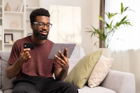 Téléchargez les photos : Homme afro-américain souriant faisant du shopping en ligne avec carte de crédit et tablette assis sur un canapé. Concept de commerce électronique et de paiement en ligne. Focus sélectif, Espace de copie. - en image libre de droit