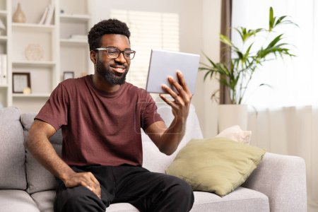 Téléchargez les photos : Heureux homme afro-américain utilisant tablette sur canapé à la maison. Homme souriant engagé dans l'appel vidéo, Loisirs Tech Lifestyle. Concept de vie confortable et de connectivité moderne. - en image libre de droit
