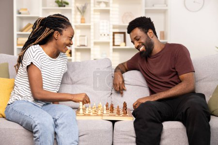 Foto de Pareja feliz disfrutando de una partida de ajedrez en casa. Hombre y mujer afroamericanos sonrientes involucrados en un partido de ajedrez amistoso, experimentando alegría y ocio en un ambiente cómodo de sala de estar. - Imagen libre de derechos