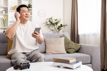 Happy Man Enjoying Music At Home, Asian Adult Smiling Using Smartphone and Headphones in Modern Living Room, Casual Clothing, Comfort, Leisure, Positive Vibes, Home Entertainment Concept