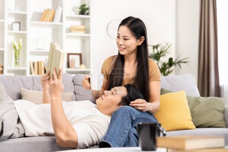 Foto de Feliz pareja asiática relajándose en casa, hombre leyendo libro mientras mujer sosteniendo taza, acogedor fin de semana, tiempo libre de tecnología de ocio - Imagen libre de derechos