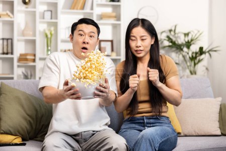 Photo for Asian couple watching a movie together, sharing popcorn and enjoying quality time on a cozy sofa. - Royalty Free Image