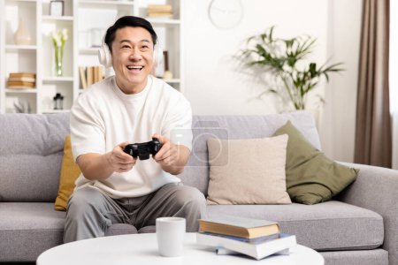 Glücklicher asiatischer Mann, der Videospiele auf der Couch spielt, die Freizeit im Haus genießt, ein unterhaltsames Unterhaltungskonzept für zu Hause entwickelt und mit Kopfhörern und Joystick spielt