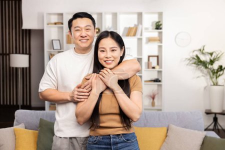 Téléchargez les photos : Couple heureux souriant à la maison : Homme et femme asiatiques dans l'usure décontractée, embrassant dans un salon confortable, exprimant l'amour et la convivialité dans un intérieur de maison moderne. - en image libre de droit