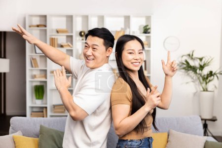 Téléchargez les photos : Couple asiatique joyeux geste heureux vers l'espace vide dans un salon moderne confortable, indiquant un objet invisible. - en image libre de droit