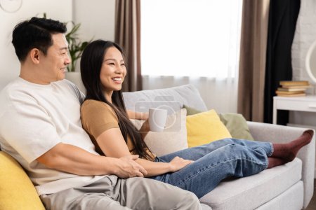 A happy Asian couple is relaxing on the sofa at home, enjoying a cozy moment with laughter and comfort.