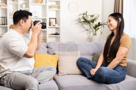Foto de Un hogar alegre captura a un hombre tomando fotografías de una mujer sonriente sentada cómodamente en un sofá. - Imagen libre de derechos