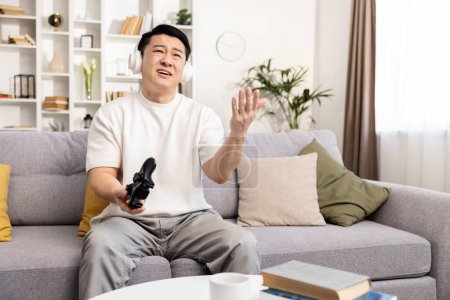 Téléchargez les photos : Un homme adulte semble frustré en jouant à des jeux vidéo, assis sur un canapé à la maison, portant un casque et tenant une manette de console. - en image libre de droit