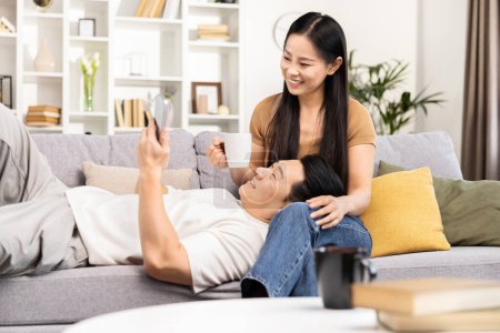 Photo for Happy Asian couple relaxing at home, man lying on couch using smartphone with smiling woman beside him holding a coffee mug. - Royalty Free Image
