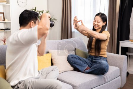 Ein junger Mann fotografiert eine Frau, die es sich in einem gemütlichen Wohnzimmer auf einem Sofa bequem macht.