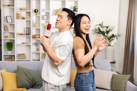 Foto de Joyful pareja asiática teniendo una divertida sesión de karaoke en casa. El hombre canta en un micrófono mientras la mujer aplaude y aplaude. - Imagen libre de derechos