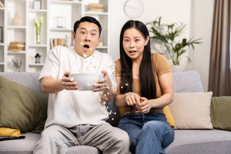 Foto de Pareja asiática en el sofá viendo una emocionante película juntos, con palomitas de maíz derramándose en un momento de sorpresa. - Imagen libre de derechos