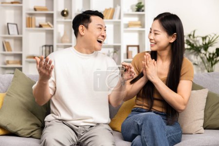 Téléchargez les photos : Couple heureux appréciant la conversation sur le canapé, homme et femme asiatiques riant et gestualisant, environnement familial détendu, interaction joyeuse, salon confortable, convivialité familiale, émotions positives - en image libre de droit