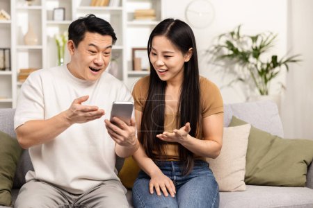 Téléchargez les photos : Couple asiatique excité Découvrir contenu amusant sur Smartphone à la maison. Capture franche capturant la joie, la surprise et l'utilisation de la technologie dans la vie quotidienne. - en image libre de droit