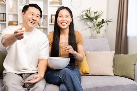 Una pareja relajada se sienta en su cómodo sofá, viendo televisión y meriendas en un acogedor ambiente de sala de estar.